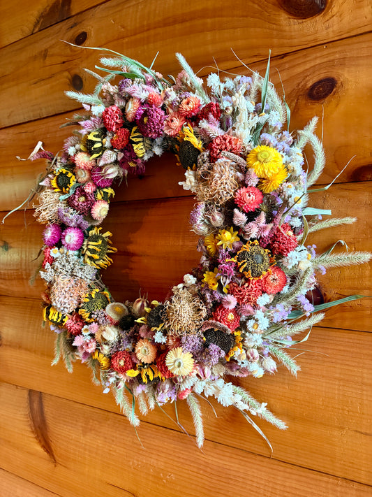 A rustic dried flower wreath with sunflowers, pink and purple blossoms, and green grasses, displayed on a warm wood plank wall.