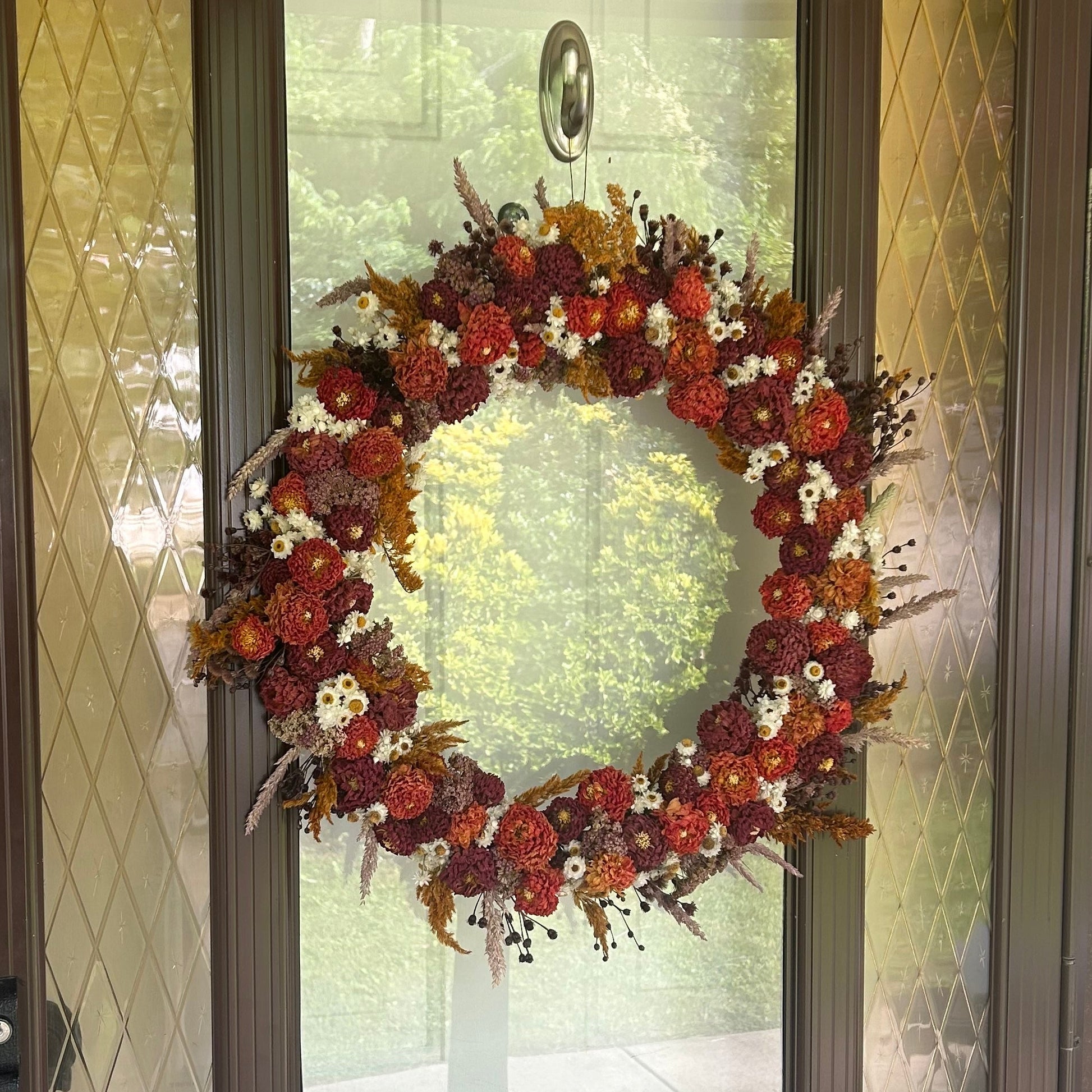 An autumn-themed wreath made of red berries, orange pinecones, and white flowers hanging on a brown glass-front door.