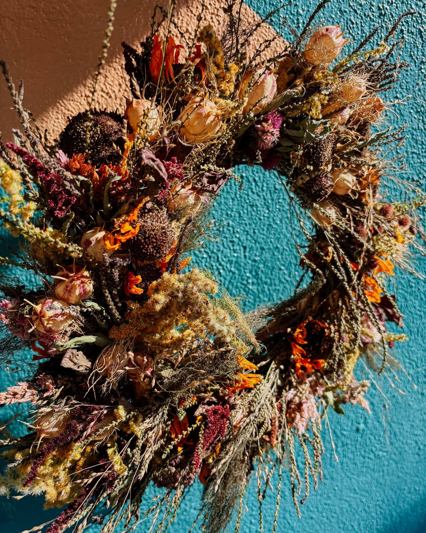 A rustic dried flower wreath with orange, purple, and beige botanicals, hanging against a textured teal wall.