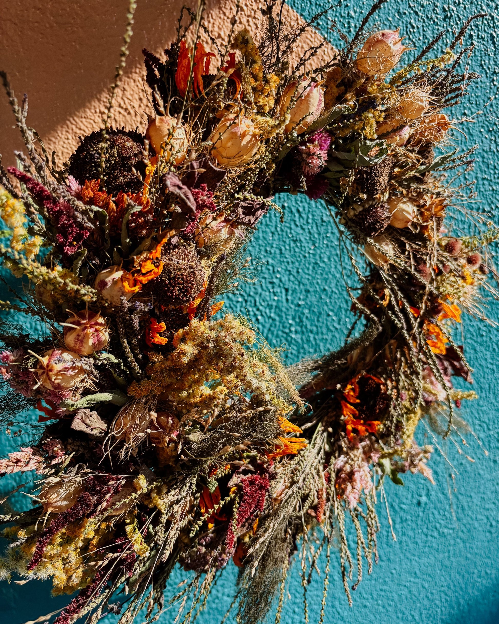 A rustic dried flower wreath with orange, purple, and beige botanicals, hanging against a textured teal wall.